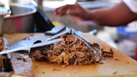 Close up of a Mexican food stand, where a serving of juicy birria (stewed beef) is being chopped with a large butcher knife. The steaming meat is tossed with prongs as it is cut up.
