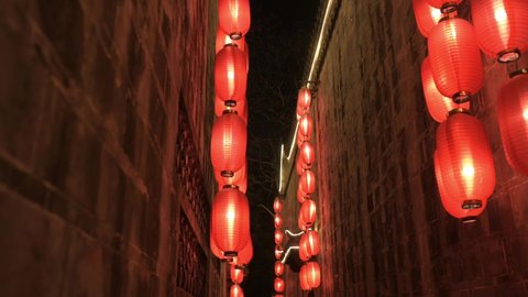 Moving forward next to right rock wall through the narrow corridor of old Chinese temple at night. Ignited red lanterns hanging around illuminating light. Traditional decoration, holiday celebrations