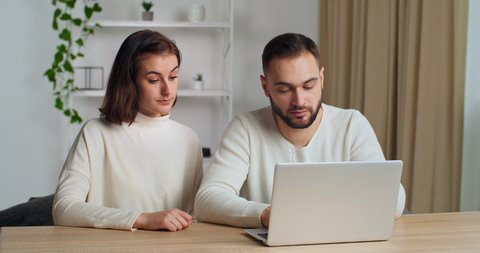 Caucasian couple 30s years old wife and bearded husband in living room using laptop enjoy e-shopping remotely, planning future relocation choose renovation firm services online. E-commerce concept
