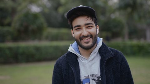 Static shot of a cheerful young Indian handsome man wearing a cap and a sports jacket with a broad smile on the face standing in a public park looking at the camera.