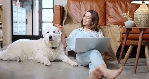 Young woman works on a laptop while sitting with her adorable white dog on the floor at cozy home