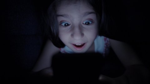 Beautiful little girl spends time with tablet computer under the duvet in the dark. Playing online games. Internet or online game addict concept.