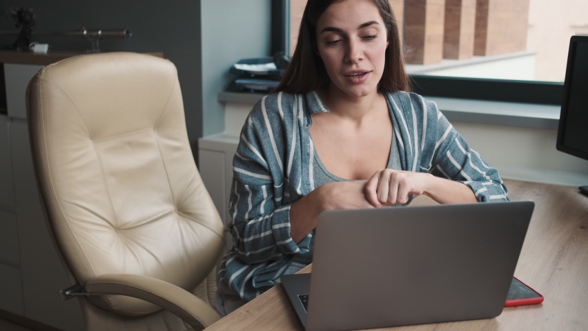 A concentrated young woman is talking with video connection using her laptop computer while sitting in the office | Shutterstock HD Video #1068093380