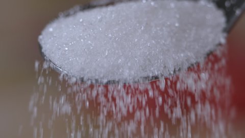 Slow Motion Close Up Locked Down Shot of White Sugar Crystals Falling Off Spoon. Closeup Macro of Pouring Full Spoon Crystals White Sugar Slow Motion. Unhealthy Diet, Diabetes, Obesity.