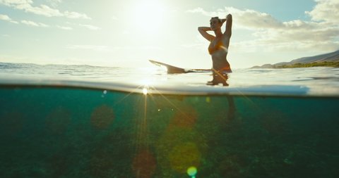 Beautiful female surfer sitting on surfboard in ocean at sunset waiting for the next wave. Slow motion shot on RED cinema camera.