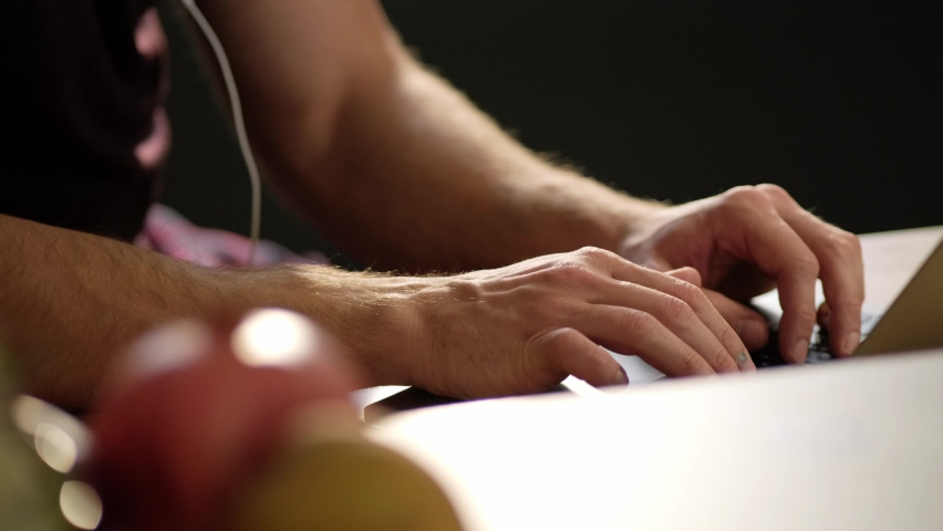 A close-up view of a young man is working with his laptop computer typing on the keyboard sitting in the apartments