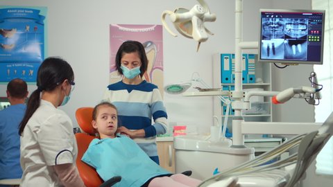 Dentist technician with gloves explaining the process to kid patient before surgery lighting the lamp. Orthodontist speaking to girl with toothache sitting on stomatological chair.