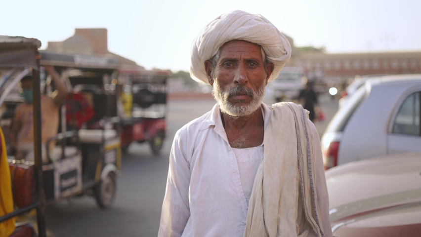 Frontal close slow motion shot of an old Indian male villager in traditional attire with a turban on head standing next to a busy road in a market place and looking at the camera