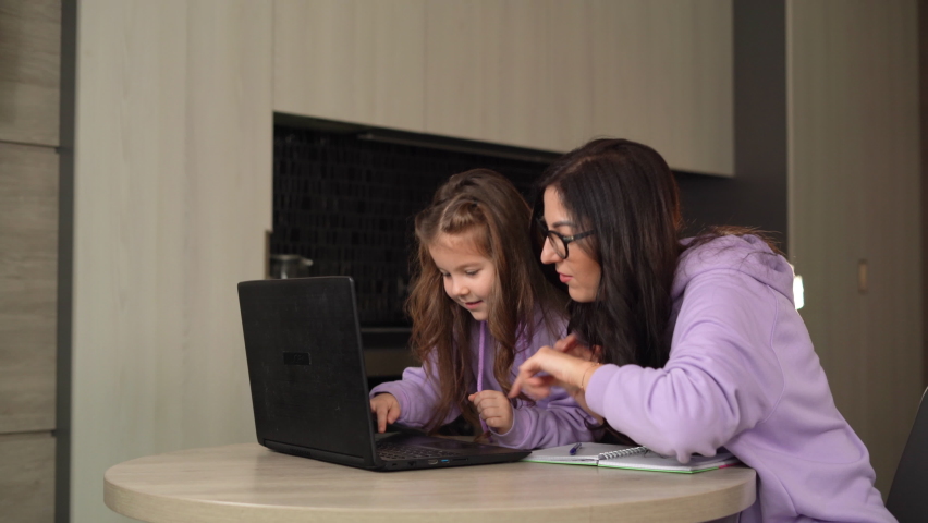 Mom and daughter are sitting together at home in the kitchen at the table. A woman teaches a child to use the Internet and a laptop. | Shutterstock HD Video #1068105647