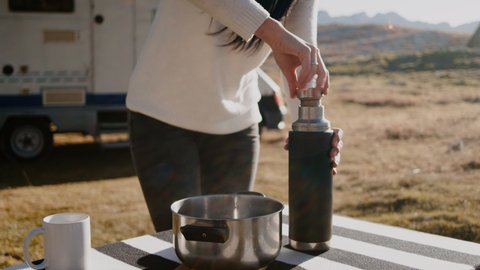 Woman using thermos and standing at table while traveling by camper car in countryside rbbro. Closeup view of young female traveler closes bottle cap and stands at desk outdoors on autumn day, man is