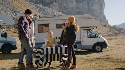 Family eat by table on motorhome road trip. Rbbro Children and parents talk and have lunch outdoor in mountains, enjoy food drink and nature. Camper RV. Mother and father kiss, kids son and daughter