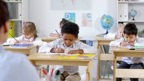 Medium shot of cute little African-American boy sitting at his desk and writing in exercise book, then raising hand and answering question asked by female primary teacher