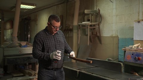 master man in shirt, jeans and gloves covers wooden piece with solution, varnish, paint, protective emulsion from spray, close-up spray, production of furniture, wooden objects, workshop, production
