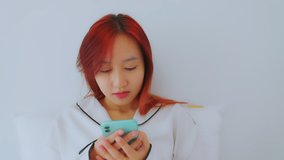 A beautiful asian woman red hair dye color in white pajamas on bed. She play social media on mobile phone in the morning on holiday. Relax, smile happily. Lifestyle Concept.