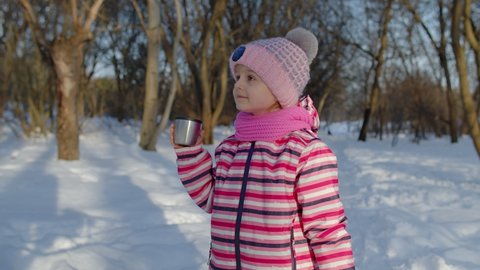Smiling child kid drinking hot drink tea from cup, walking in winter park forest. Portrait of little girl trying to keep warm outdoors in frosty day sunset. Children at Christmas holidays eve time