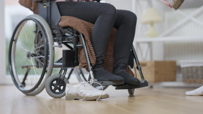 Caring young Caucasian woman putting shoes on paralyzed legs of Person with a disability sitting using a wheelchair. Volunteer or friend helping woman with paraplegia indoors. Care and disability concept. Royalty-Free Stock Footage #1068114716