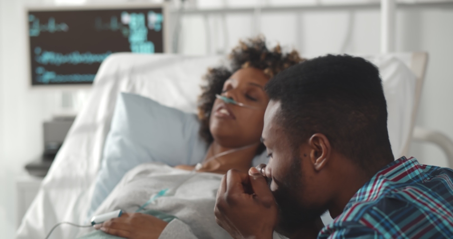 Worried african husband sitting at sick wife bed in hospital. Young afro-american woman patient sleeping in bed at clinic ward with sad husband sitting at bedside | Shutterstock HD Video #1068116090