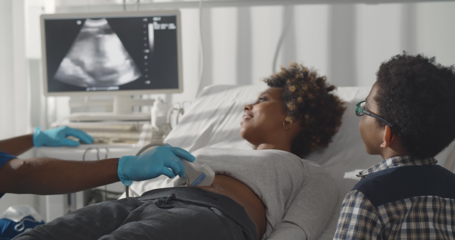 Pregnant african woman with elder son visiting gynecologist. Afro-american kid looking at screen while doctor examining mother and checking belly with ultrasound Royalty-Free Stock Footage #1068116123
