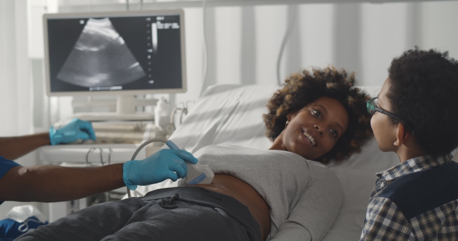 Pregnant african woman with elder son visiting gynecologist. Afro-american kid looking at screen while doctor examining mother and checking belly with ultrasound