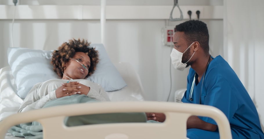 African young doctor in mask consulting covid-19 infected patient in bed at hospital. Sick black woman lying in hospital bed and talking to surgeon in scrubs and protective mask Royalty-Free Stock Footage #1068116267