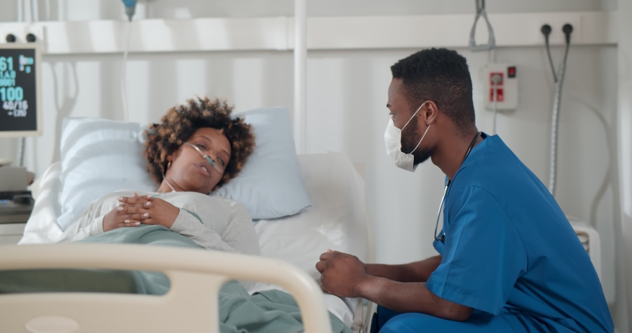 African young doctor in mask consulting covid-19 infected patient in bed at hospital. Sick black woman lying in hospital bed and talking to surgeon in scrubs and protective mask | Shutterstock HD Video #1068116267