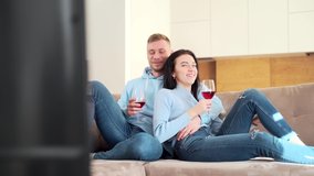 young cheerful couple watching funny movie on tv at home. A man and a woman or a boyfriend and a girlfriend are sitting on the couch with a glass of wine watching a TV series and laughing.