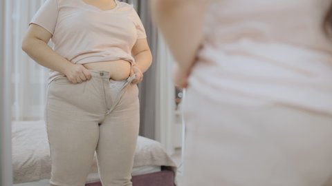 Obese female trying to zip up tight trousers in front of mirror, plus size