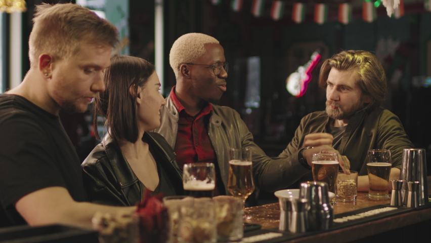 Waist-up footage of happy young group of multi-ethnic friends having fun together sitting at bar drinking beer and chatting