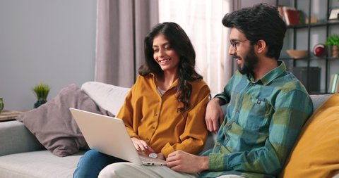 Portrait of young Indian happy cheerful family couple resting on sofa in living room searching internet typing online on laptop computer and speaking, choosing decor, e-commerce, leisure concept