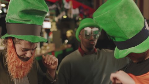 Medium close up of multi-ethnic male friends in Irish costumes having fun together crazy dancing in local pub, celebrating St Patricks Day