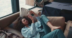 Cinematic shot of happy young beautiful mixed race woman lying on sofa at home, playing smartphone game and smiling.