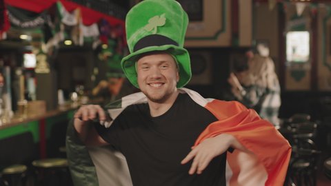 Medium close up portrait with slowmo of energetic caucasian man dancing to camera with Irish flag on his shoulders doing wave moves, celebrating St Patricks Day in pub with friends
