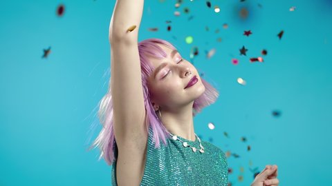 Blissful trendy woman dancing, touching unusual violet dyed hair during confetti rain in blue studio. Concept of celebrating, party, winning