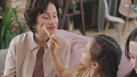 High-angle slow-motion close up with tracking of cheerful little girl holding make-up brush, applying blush on mom and grandma cheeks, having fun together sitting on couch in living room