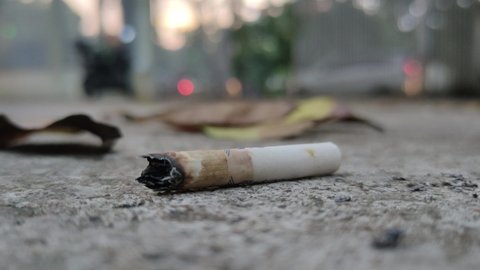 Close up a cigarette butt on the ground with evening traffic, fallen leaves and the feet of a woman walking background at home page. Illustration of quitting smoking. Cinematic video.