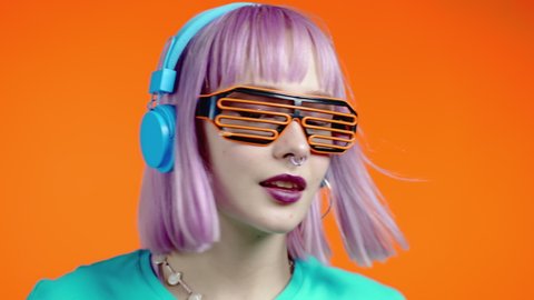 Unusual trendy woman with dyed pink hair listening music, dancing in headphones on orange background. Color block. Fashionista teen girl in lattice glasses. Slow motion.