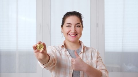 Young woman holding a gold coin of cryptocurrency in her hand and smiling against the background of a window.