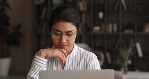 Pensive young indian businesswoman in eyeglasses looking at laptop screen, thinking on problem solution. Thoughtful beautiful mixed race employee working on difficult task online on computer.
