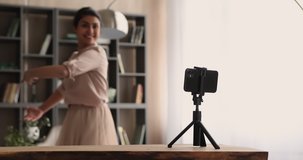 Focus on tripod with smartphone recording energetic young indian woman dancing. Happy millennial mixed race lady making classic dance ballet moves, filming video on cellphone web camera indoors.