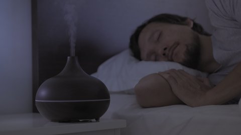 At night man slips in bed with an aromatherapy diffuser working on a nightstand. Deep and restful sleep concept