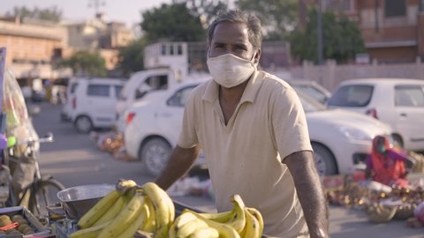 Indian middle-aged male local fresh fruit vendor wearing a protective face mask standing by a carrier cart around a corner at the busy market street looking in the camera during COVID 19 epidemic