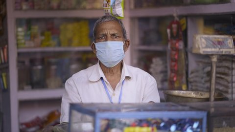 Frontal shot of an Indian Asian adult grocery shopkeeper or owner wearing a protective mask on a face sitting on a shop counter looking at camera during the hard times of COVID 19 epidemic