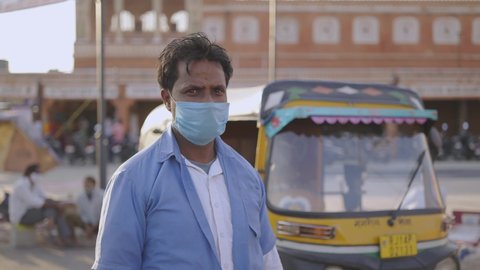 Close view shot of a young Indian auto-rickshaw driver standing in front of his CNG vehicle with a protective mask on the face staring at the camera during the hard times of the COVID 19 epidemic