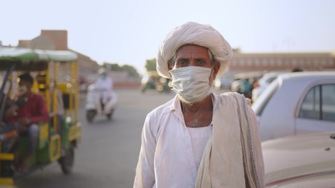 Close shot of an old Indian male villager in traditional attire wearing face protective mask and standing next to a busy road in a market place while looking at the camera amid Corona virus epidemic