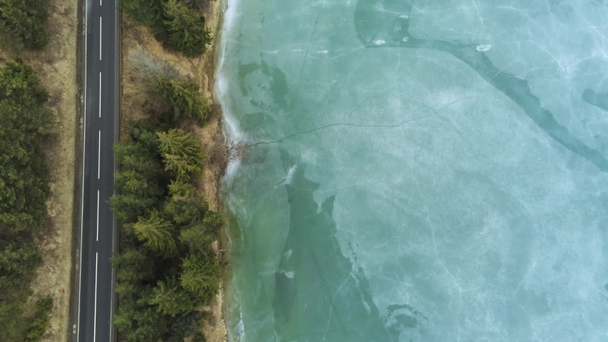 Drone camera flying over beautiful blue frozen crystal clean lake near asphalt road .