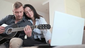 young couple learning online to play guitar while sitting at home on sofa with laptop. guy and a girl or a man and woman learn a musical instrument from video lessons distance video tutorial studied