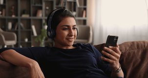 Happy beautiful young woman in wireless headphones holding mobile video call conversation with friends, talking chatting discussing life news distantly, resting on comfortable couch at home.