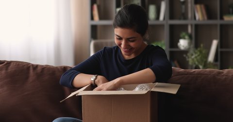 Curious happy young beautiful indian ethnicity woman sitting on sofa, unpacking carton box, feeling satisfied with fast delivery service or ordered item, having positive online shopping experience.