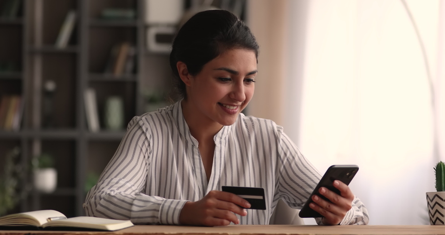 Happy millennial indian ethnicity woman holding bank card, entering cvv code or payment information in mobile internet shopping application, feeling amazed of purchasing goods or services online. Royalty-Free Stock Footage #1068157376
