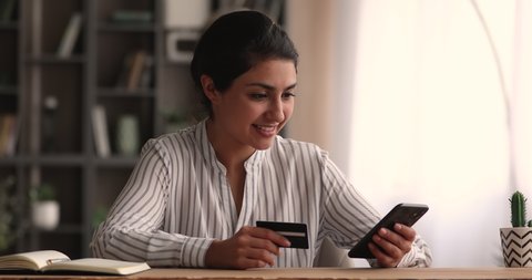 Happy millennial indian ethnicity woman holding bank card, entering cvv code or payment information in mobile internet shopping application, feeling amazed of purchasing goods or services online.